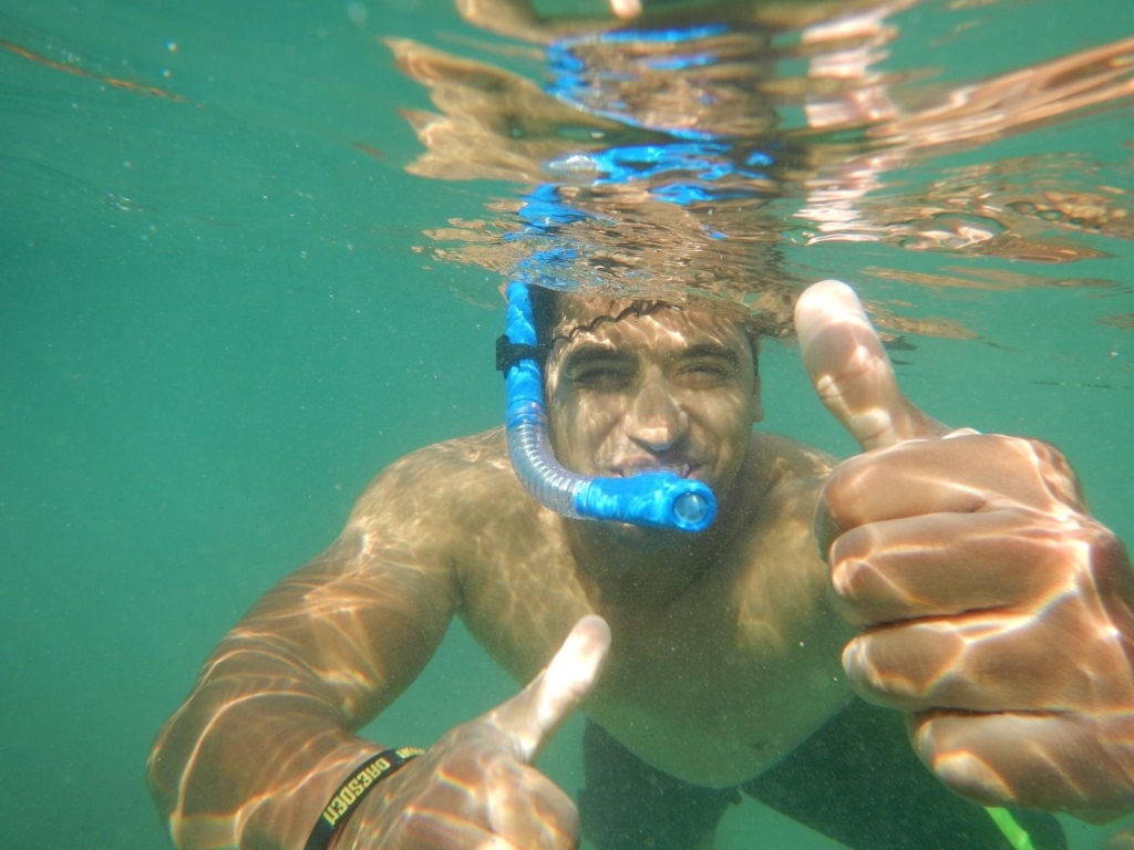 An Asian man wearing a blue snorkel under water giving a thumbs up. 